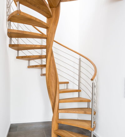 Spindeltreppe in der Holzart Eiche geölt mit durchgehendem Krümmling. Innenliegend eingestemmte und freiseitig freitragend. Geschweifte Stufenvorderkanten und ein Relinggeländer Milano 5.