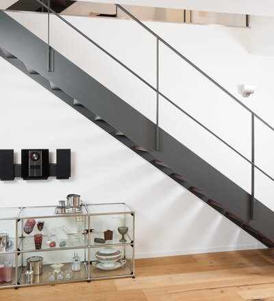 Gerade Treppe mit unsichtbar befestigten Holzstufen. Das Geländer ist aus filigranem Flachstahl hergestellt und vermittelt durch nur einen Zwischengurt eine besondere Leichtigkeit. Bei der Anordnung des Zwischengurtes wurde der „Goldene Schnitt“ angewendet. Dieser stammt aus der griechischen Antike und gibt dem Auge des Betrachters ein harmonisches Teilungsverhältnis.